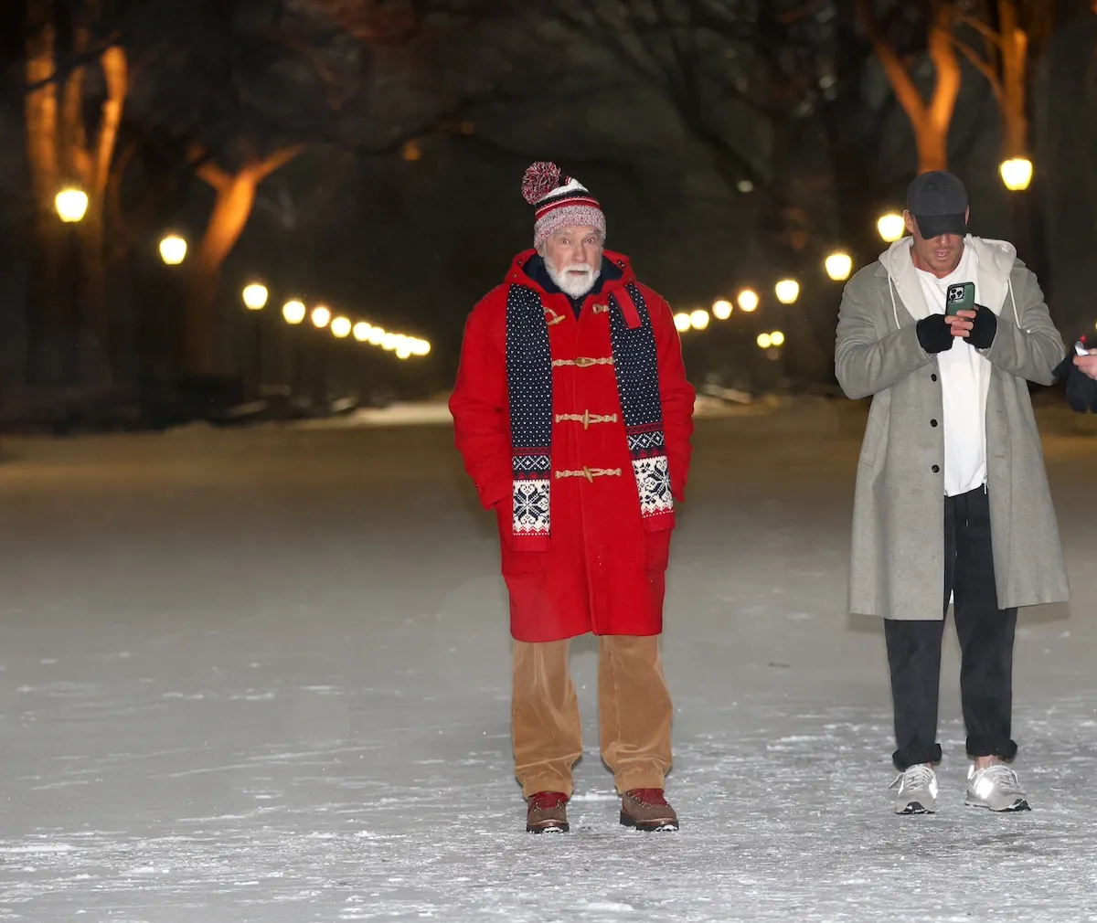 Arnold Schwarzenegger, wearing a red coat, stands in the snow while Alan Ritchson looks at his phone