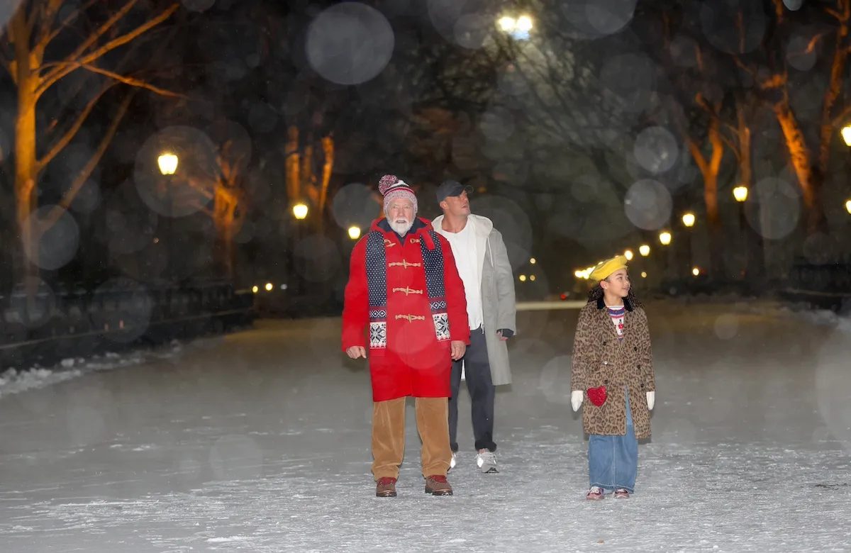 Arnold Schwarzenegger, in a red coat, stands next to Alan Ritchson and Berkeley James in a snowy Central Park