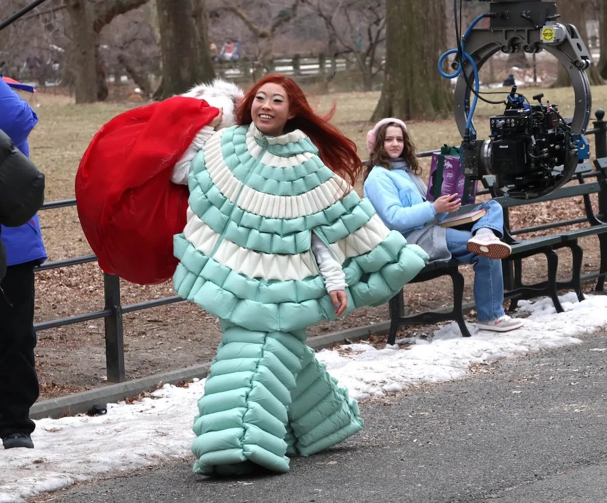 Awkwafina, dressed in a puffy blue outfit and carrying a big red sack, walks through Central Park