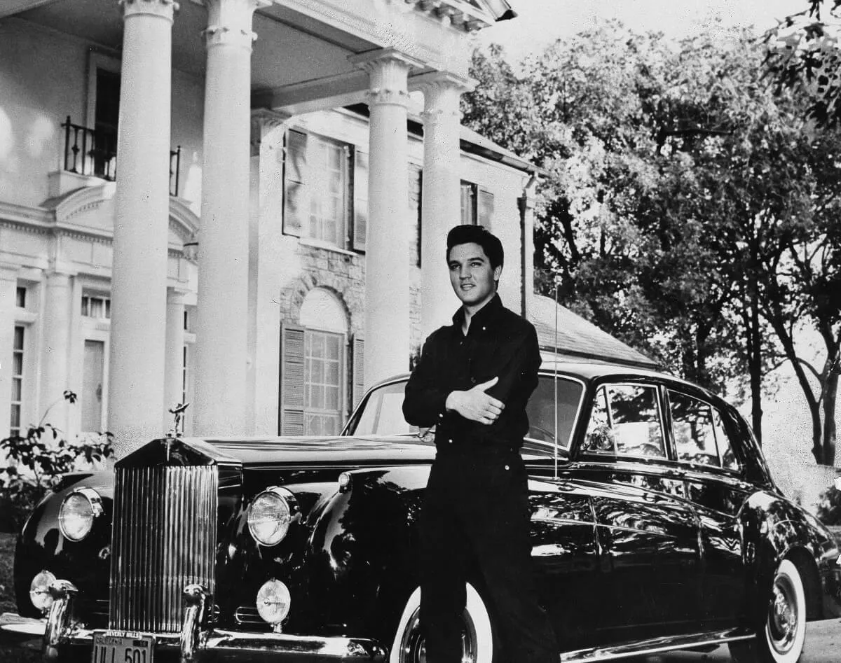 A black and white picture of Elvis standing with his arms folded in front of Graceland and a car.