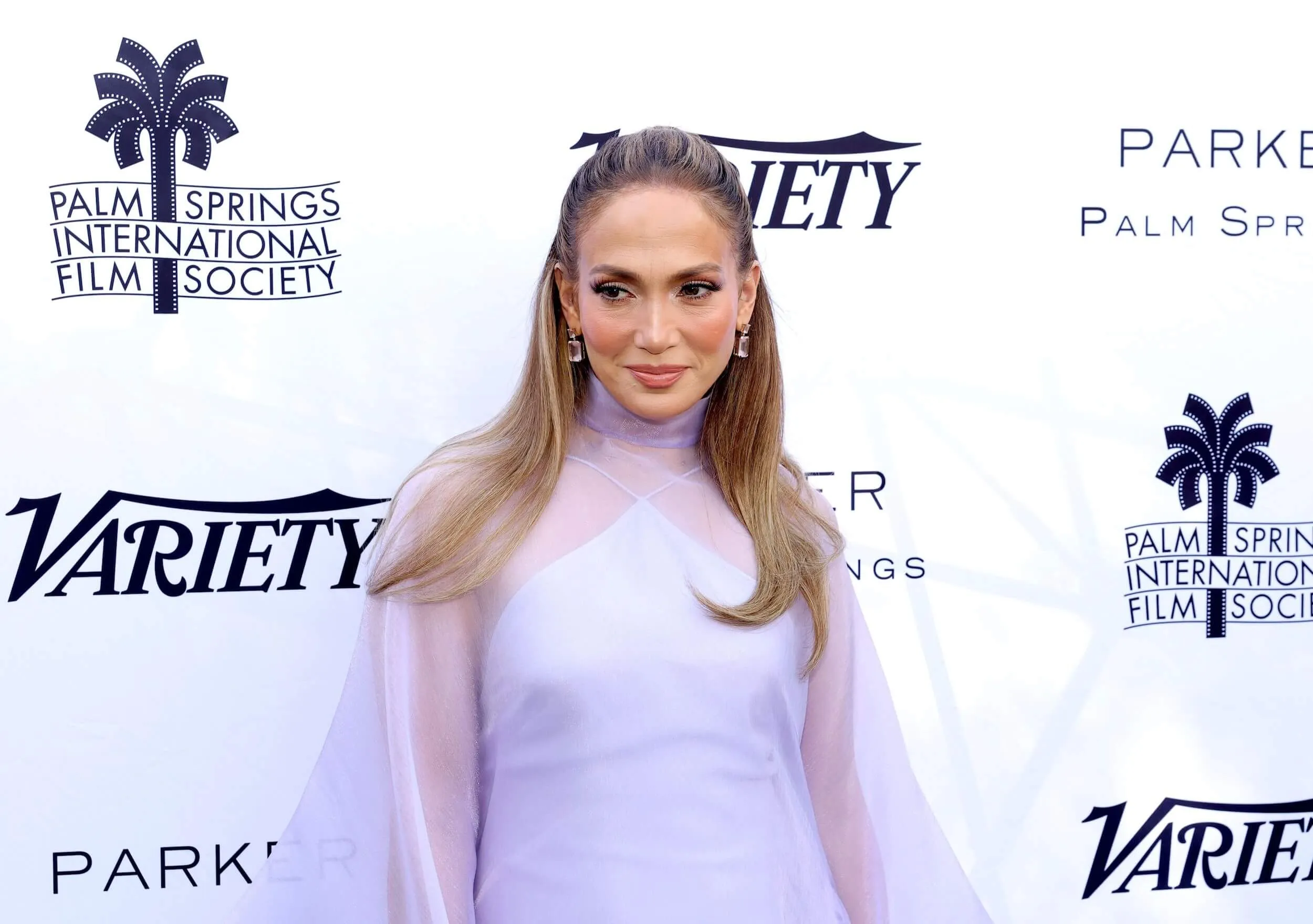 Jennifer Lopez against a Variety background. She's wearing a sheer long-sleeve dress.