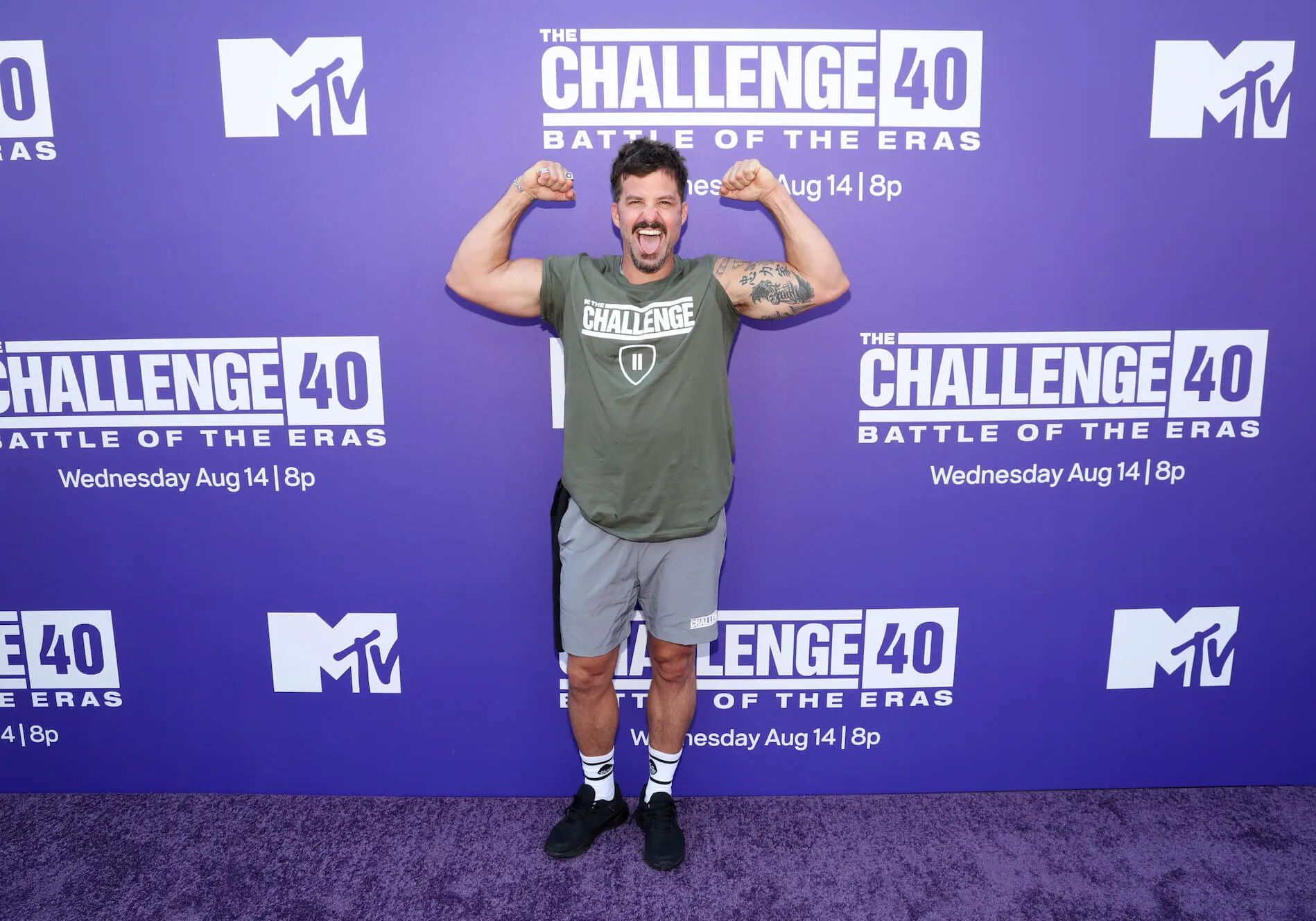 Johnny 'Bananas' Devenanzio in front of 'The Challenge 40' purple backdrop. He's posing by flexing his biceps.