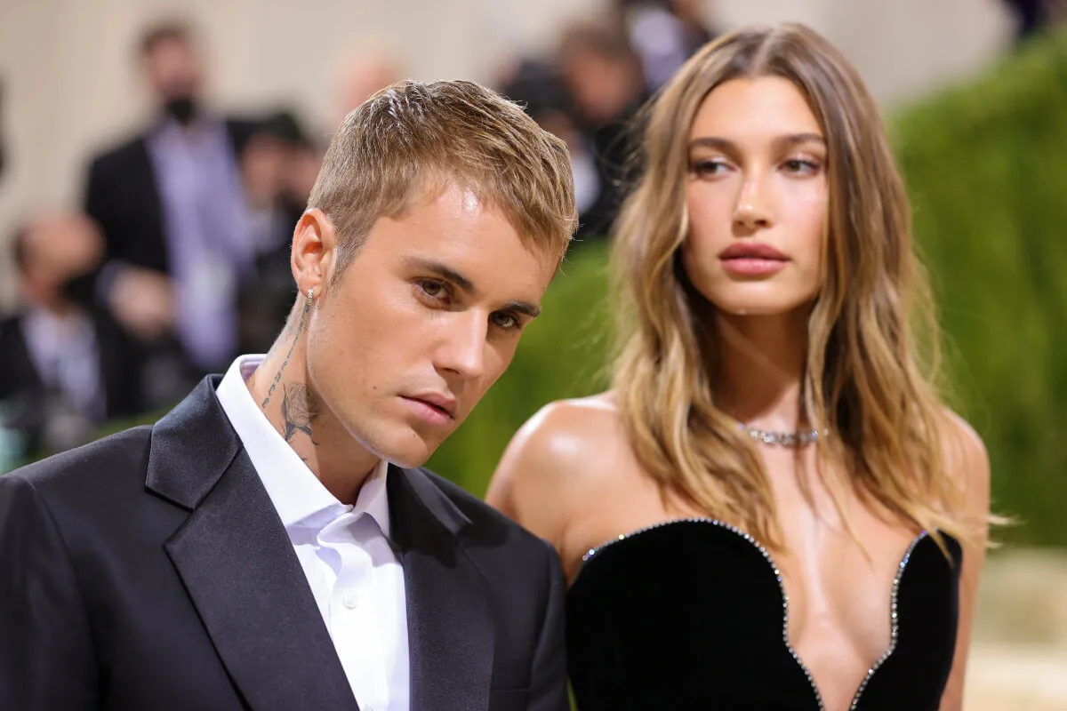 Justin Bieber stands with Hailey Bieber. He wears a suit and she wears a black strapless dress.