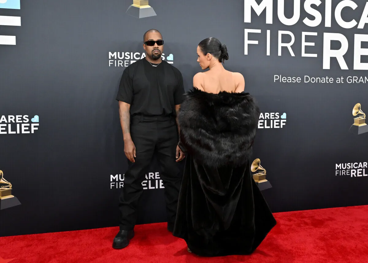 Kanye West wears a black shirt and pants with sunglasses. Bianca Censori wears a black coat and faces away from the camera. She reveals her shoulders.