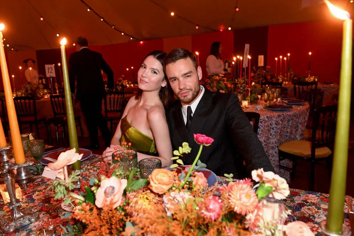 Maya Henry wears a green strapless dress and leans against Liam Payne, who wears a suit. They sit at a table covered in flowers.