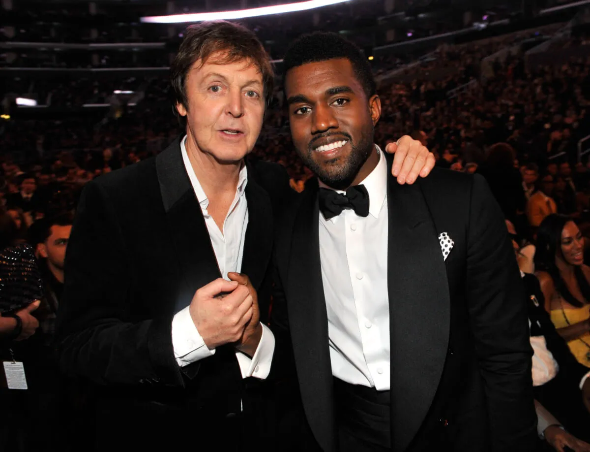 Paul McCartney and Kanye West clasp hands and McCartney has his hand on West's shoulder. McCartney wears a suit and West wears a tuxedo.