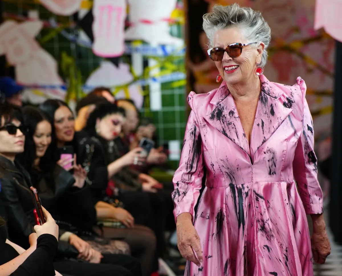 Wearing a pink satin dress, Prue Leith smiles and walks on the catwalk during the VIN + OMI London Fashion Week show